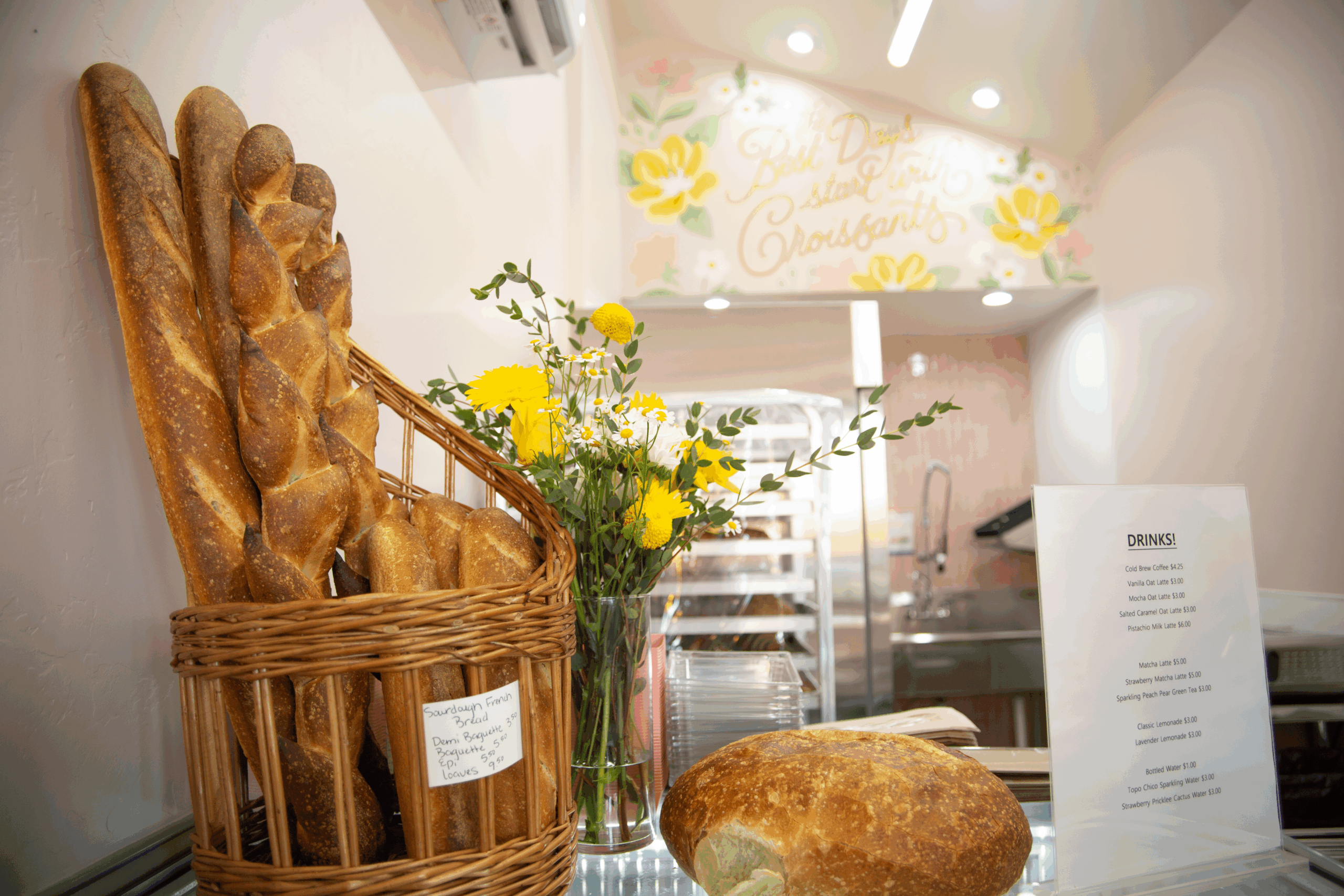 Fresh baguettes in a basket, a loaf of bread, and flowers on a counter inside a bakery